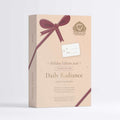 A beige rectangular gift box for The Apothecary's Daily Radiance Holiday Edition 2016, decorated with a dark burgundy ribbon graphic and a white gift tag, set against a clean white background.